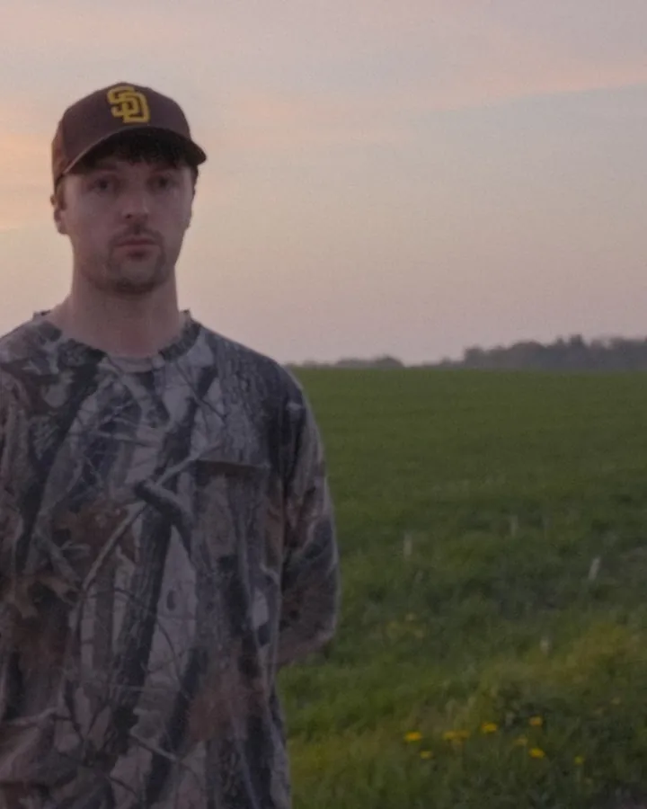 Julian Strangelove in a camo shirt and cap in a green field